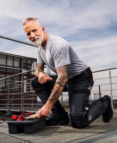 Man in geknielde positie reikt in gereedschapskist en draagt super stretch werkbroek.